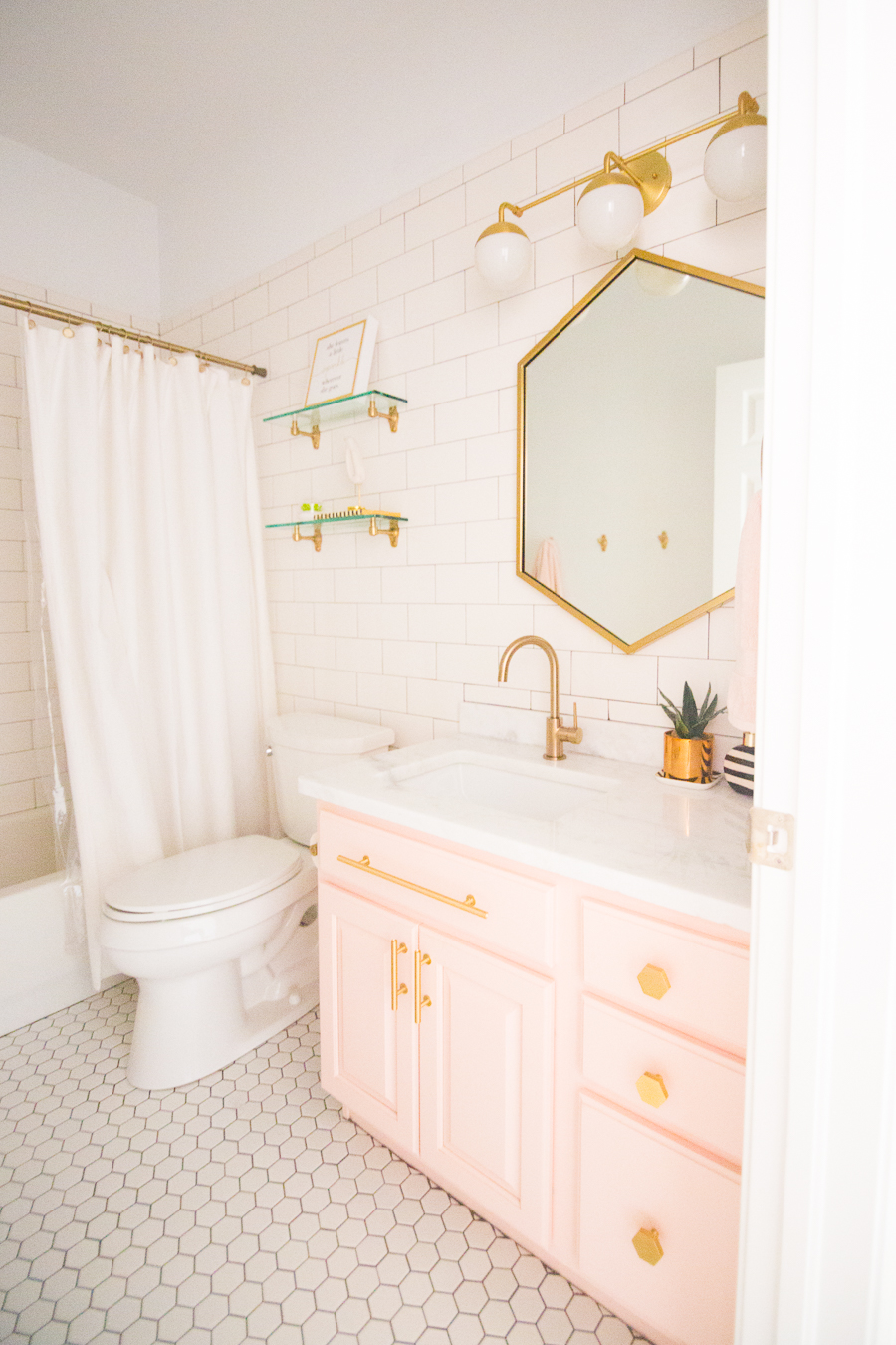 Modern Glam Blush Girls Bathroom Design gold hexagon mirror blush cabinets gold hardware white hexagon floor glass shelves pink bathroom cabinets gold orb sconce-1-3