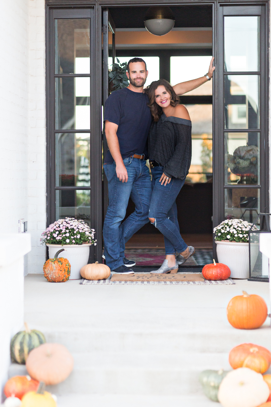 Loveliest Looks of Fall Home Tour Beautiful Fall Home Decor and Fall Fashion Ideas couple who does renovations and design sitting on the front steps of their fall front porch surrounded by pumpkins with the wife wearing old navy rockstar jeans and and off the shoulder gray top and snake skin boots and the husband wearing jeans and a navy shirt and navy cole hahn shoes