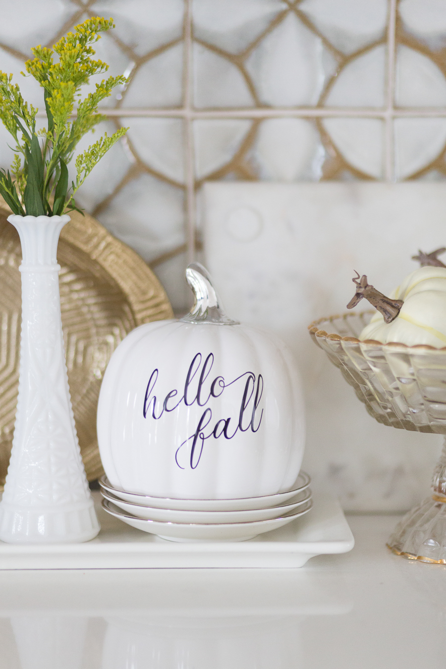 Loveliest Looks of Fall Home Tour Beautiful Fall Home Decor and Fall Fashion Ideas hello fall ceramic white pumpkin sitting on top of white plates with a cake glass dish full of white pumpkins next to it in front of patterned honeycomb ann sacks tile backsplash in a white kitchen