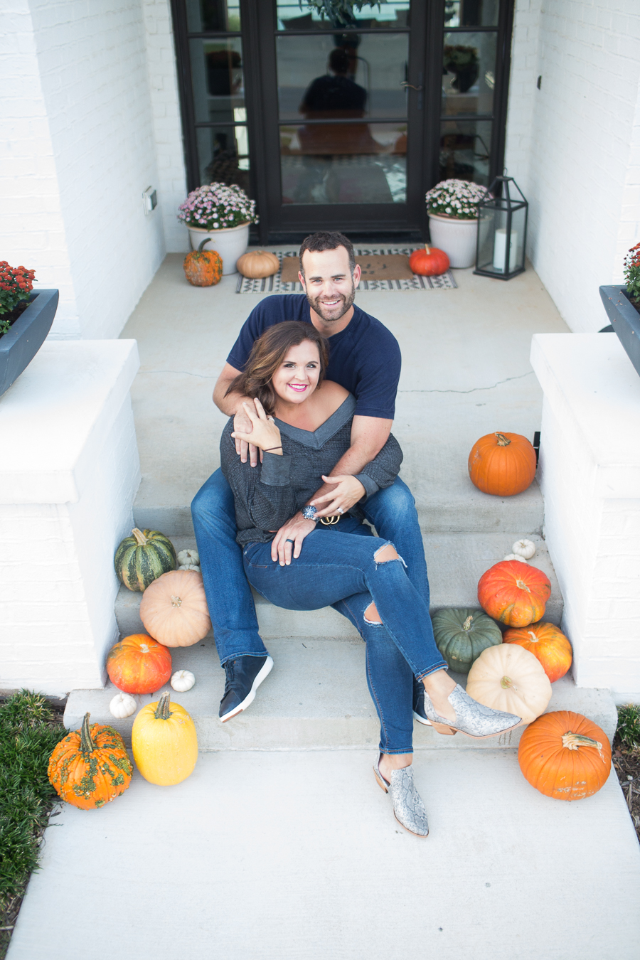 Loveliest Looks of Fall Home Tour Beautiful Fall Home Decor and Fall Fashion Ideas couple who does renovations and design sitting on the front steps of their fall front porch surrounded by pumpkins with the wife wearing old navy rockstar jeans and and off the shoulder gray top and snake skin boots and the husband wearing jeans and a navy shirt and navy cole hahn shoes