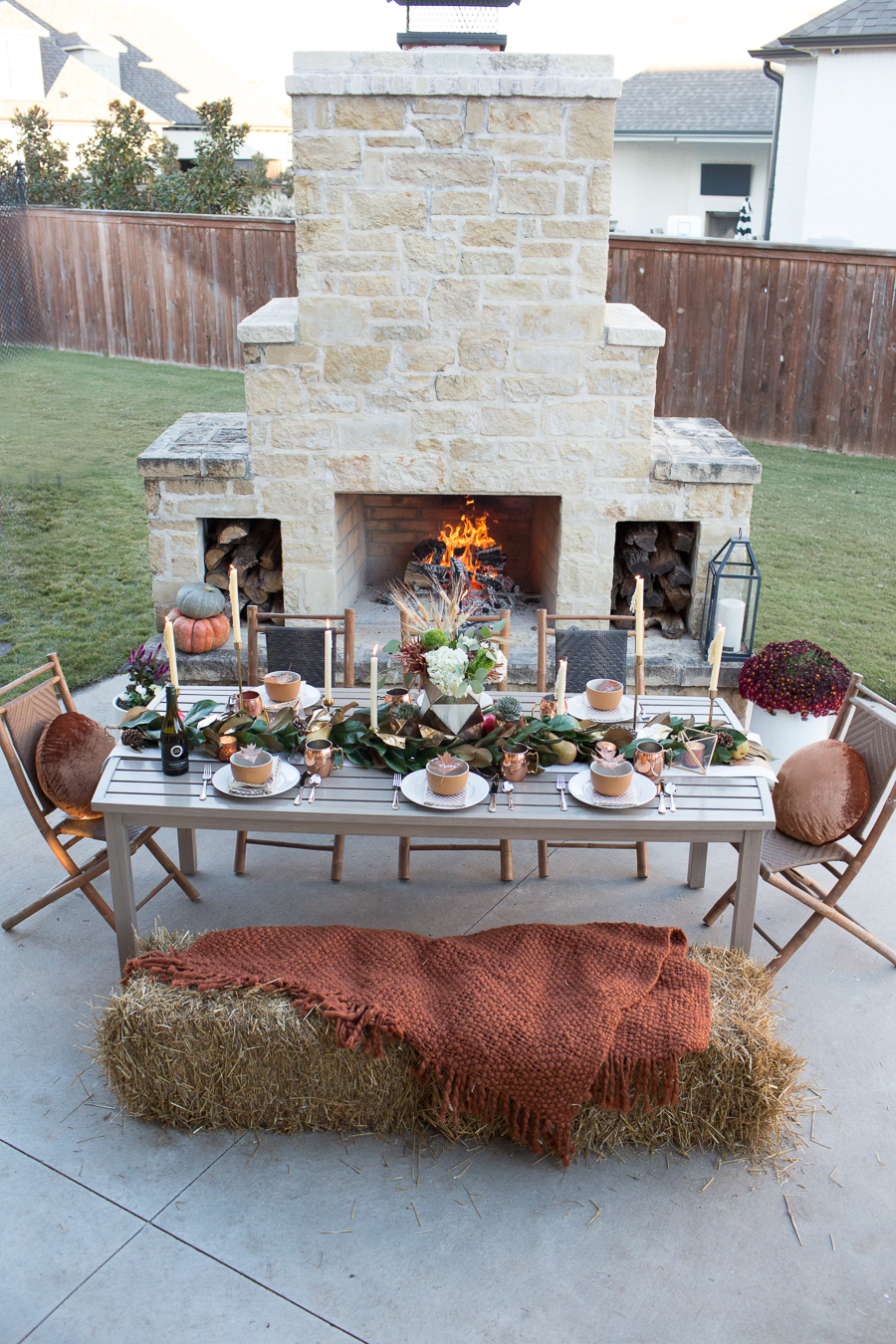 Simple Ways to Decorate for the Holidays from West Elm outdoor Friendsgiving and thanksgiving tablescape magnolia garland with gold candlesticks and a rock outdoor fireplace and copper votive candles hay bail with a chunky knit throw and copper velvet pillows