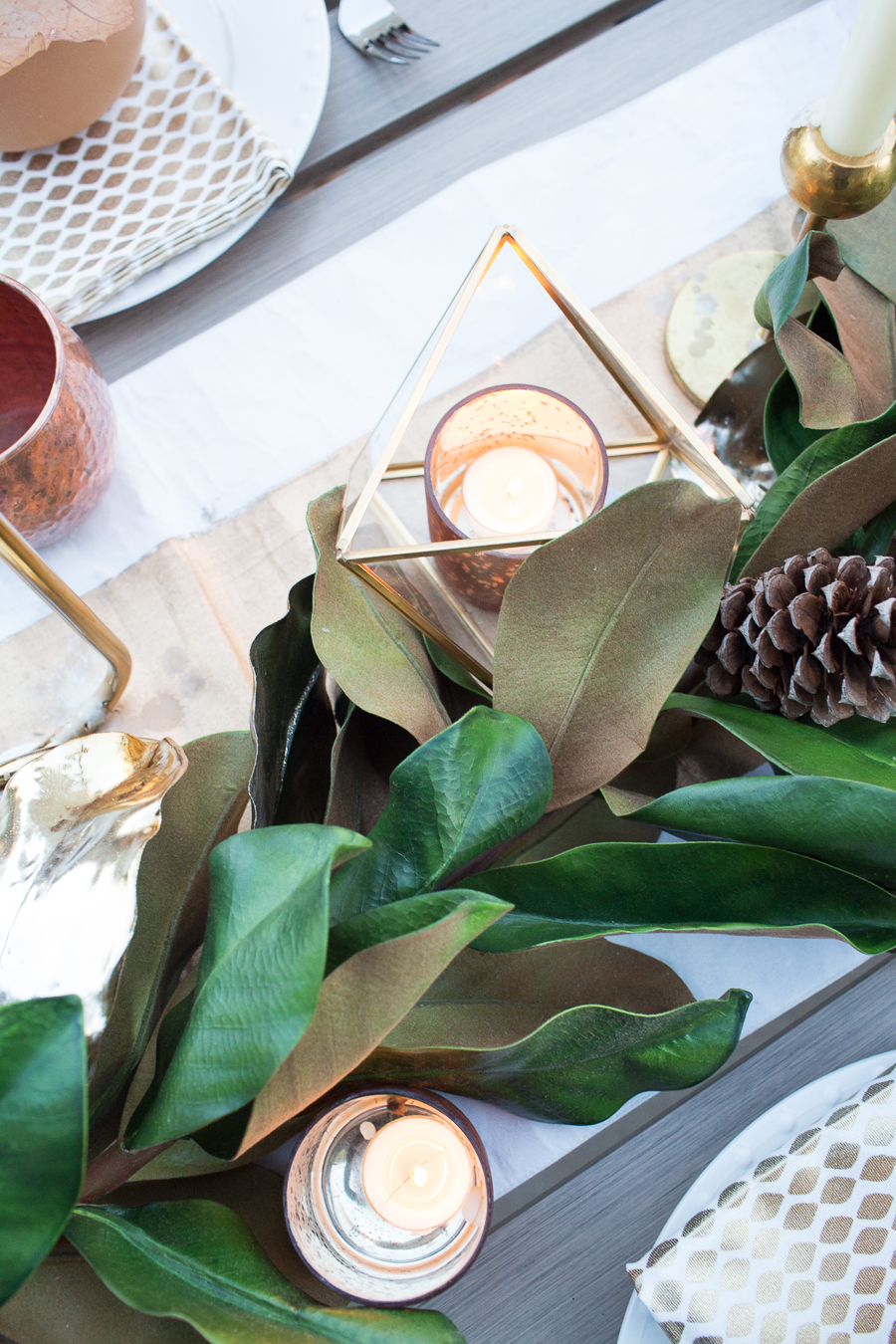 Simple Ways to Decorate for the Holidays from West Elm outdoor Friendsgiving and thanksgiving tablescape magnolia garland with gold candlesticks and a rock outdoor fireplace and copper votive candles hay bail with a chunky knit throw and copper velvet pillows