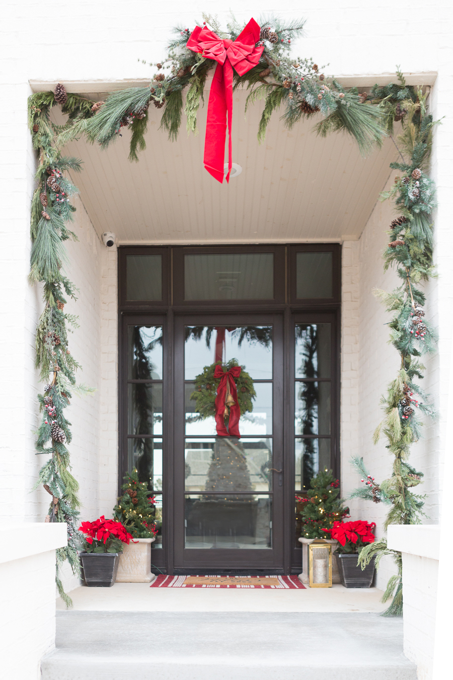 Holiday-Decor-Favorites-from-Home-Depot white brick exterior with fresh Christmas garland and red bows Christmas wreath with gold bells and a red bow silk poinsettias in a copper pot on a front porch black glass front door