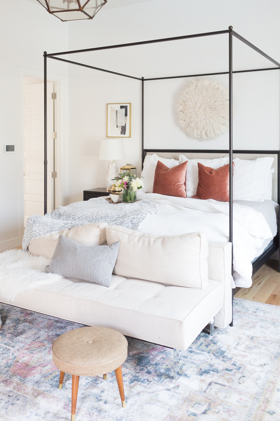 Refresh Your Master Bedroom and Bath with Pottery Barn picturing a canopy bed with white bedding, white cushioned bench at the end with tan stool and rusted colored pillows