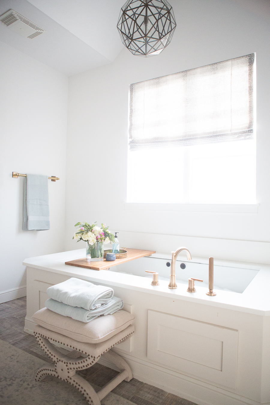 Refresh Your Master Bedroom and Bath with Pottery Barn crisp white bathroom with soaker tub, gold fixtures, a geometric light fixture, and singular window above tub