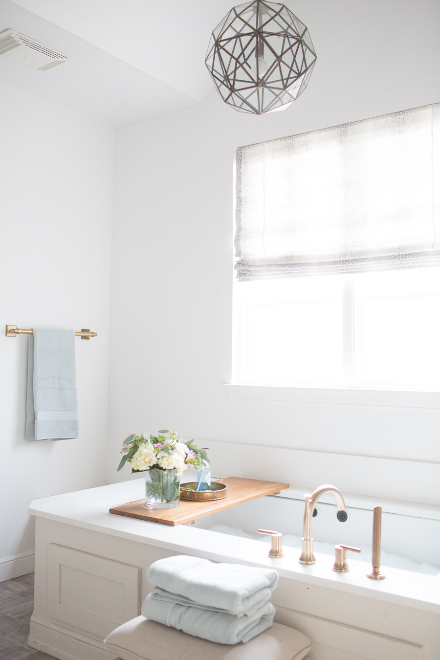 Refresh Your Master Bedroom and Bath with Pottery Barn white bathroom with geometric light fixture, soaker tub, powder blue towels and a wooden tub tray with floral arrangement on top