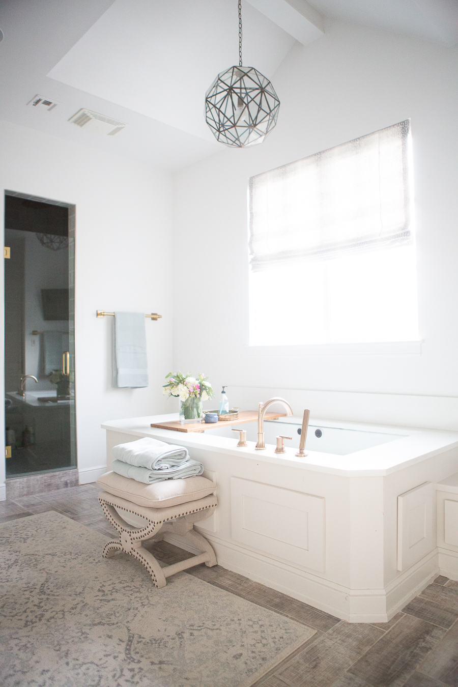 Refresh Your Master Bedroom and Bath with Pottery Barn white bathroom featuring large soaker tub, faded rug, geometric light fixture and studded stool