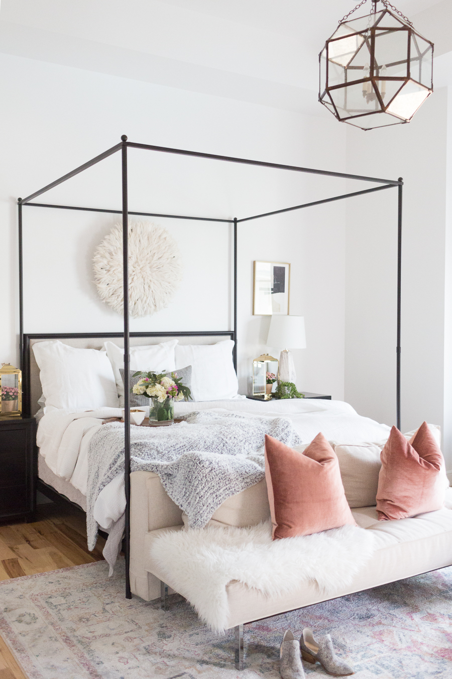 Refresh Your Master Bedroom and Bath with Pottery Barn with black canopy bed and geometric light fixture, a cream cushioned bed bench and a faded multi-colored rug