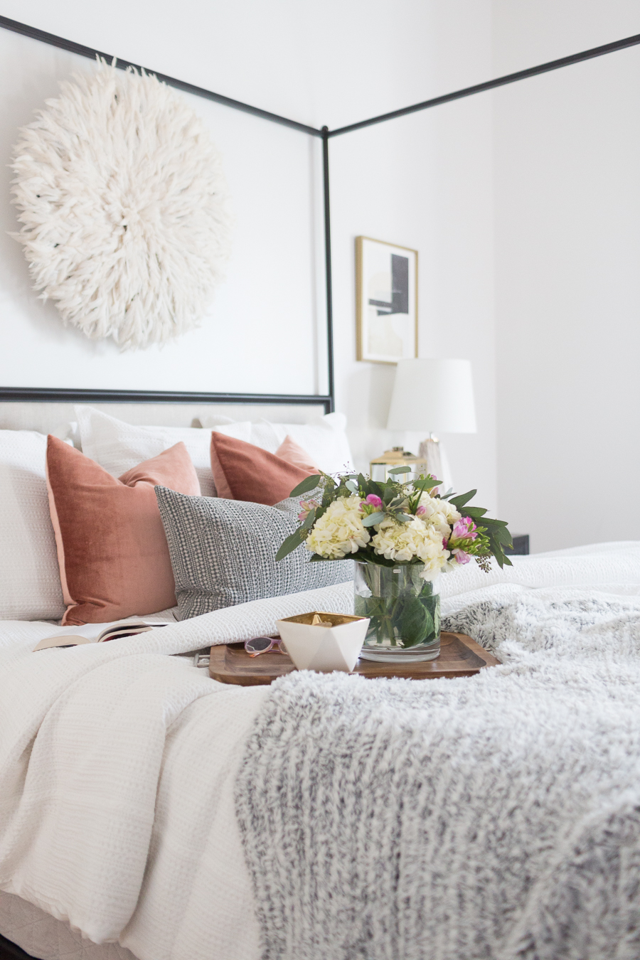 Refresh Your Master Bedroom and Bath with Pottery Barn with wooden tray sitting on top of bed with hydrangeas in a vase and white poof on the wall 