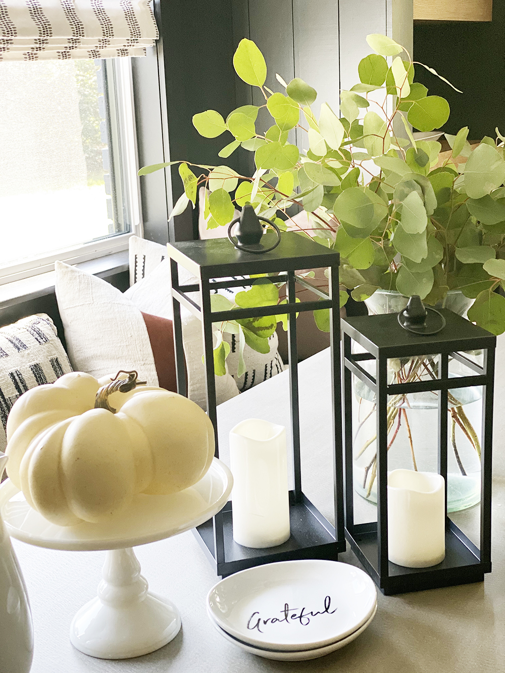 Affordable Fall Front Porch and Tabletop Decor Ideas black lanterns on a dining room table with a white cake stand and a pumpkin