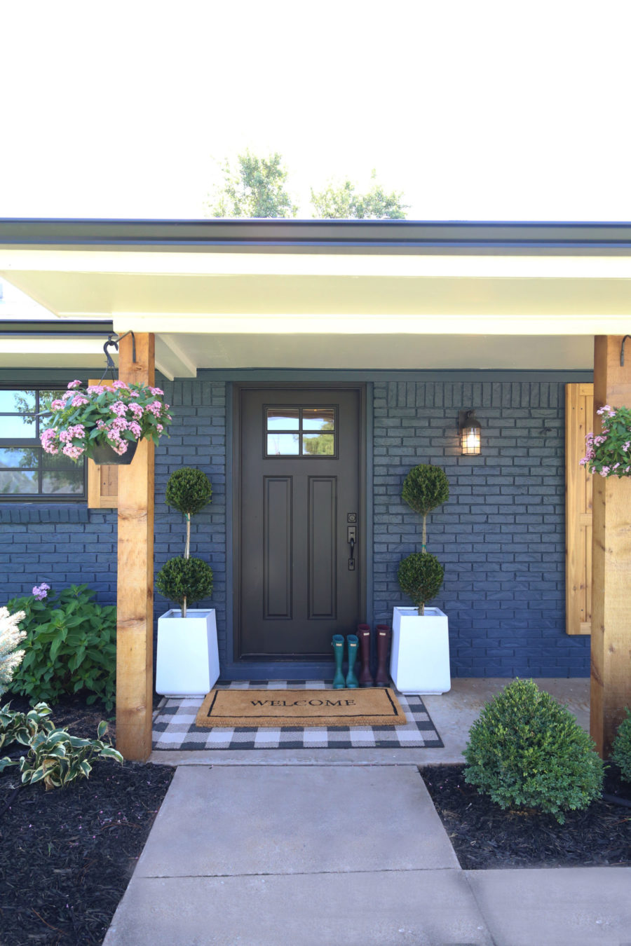 CC and Mike Modern Navy and Cedar Ranch Style Remodel navy brick with black front door and cedar posts and shutters with pink flower hanging baskets cement planters with topiaries and buffalo check rug layered on a front porch